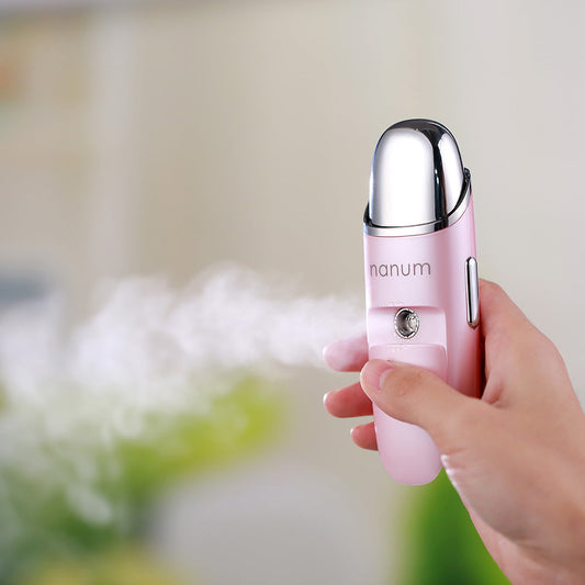Hand holding a pink portable nano spray hydrator releasing fine mist for facial hydration.